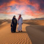 Cinematic wide-angle view of Egypt’s desert near Hurghada and Sinai, Bedouin man and woman in traditional attire with subtle facial markings, golden desert light, luxury travel editorial style, authentic heritage
