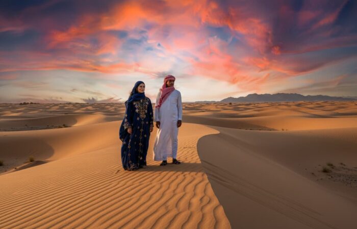 Cinematic wide-angle view of Egypt’s desert near Hurghada and Sinai, Bedouin man and woman in traditional attire with subtle facial markings, golden desert light, luxury travel editorial style, authentic heritage